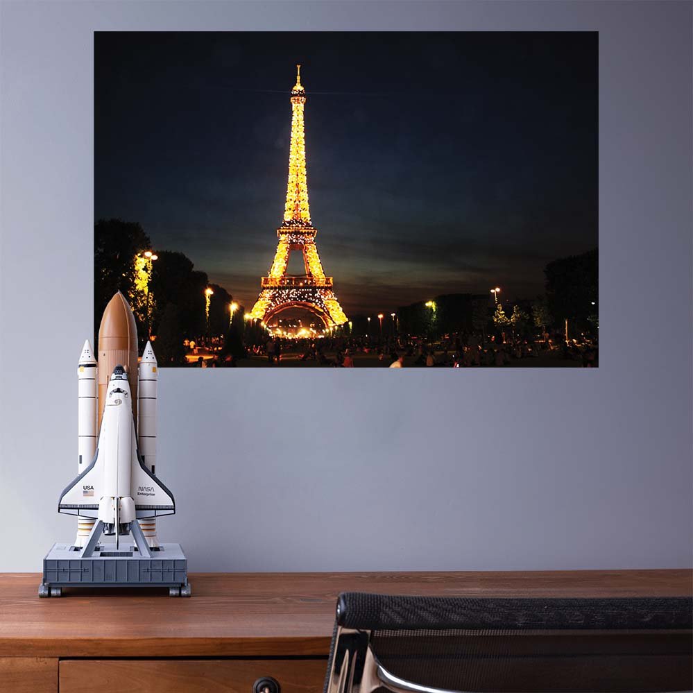 24 inch poster of the illuminated Eiffel Tower at nigh against a dark sky displayed on a wall above a desk with a space shuttle model