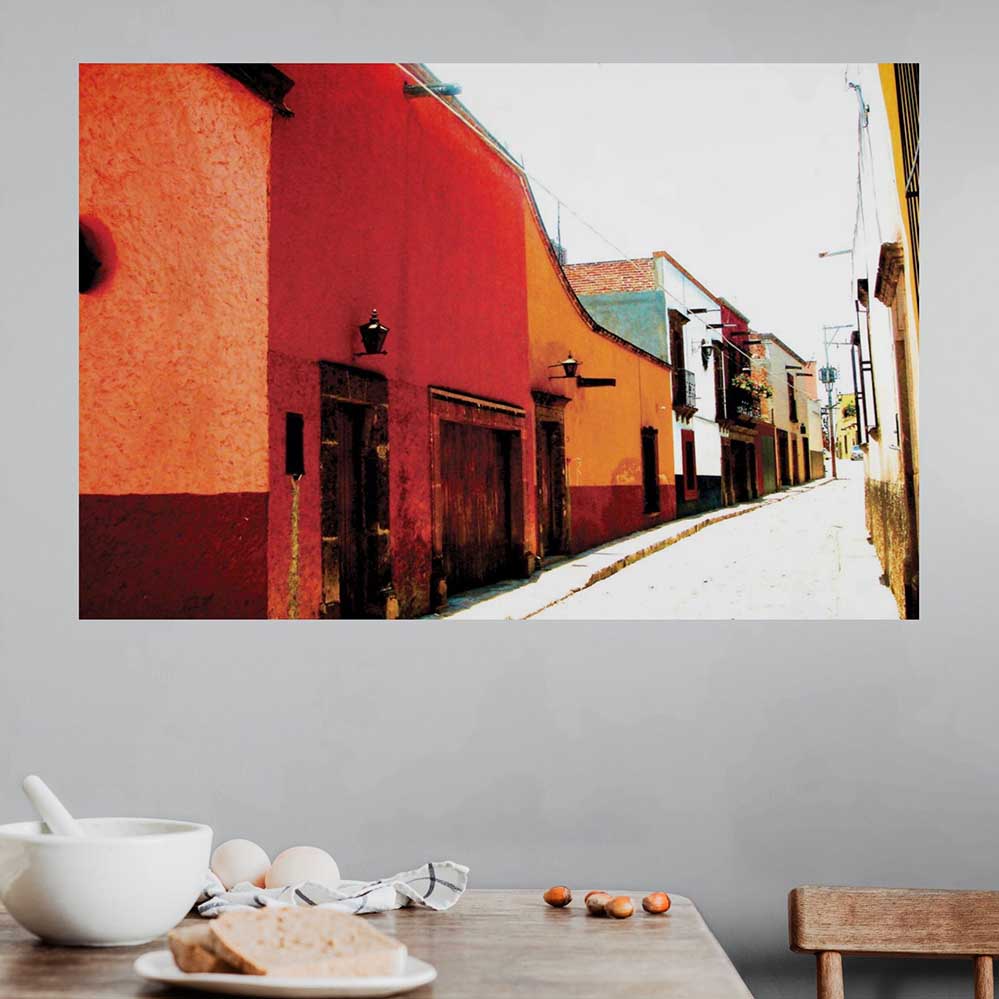 36 inch Colorful Mexican street scene poster with red and orange buildings displayed on a gray wall above a table with food.