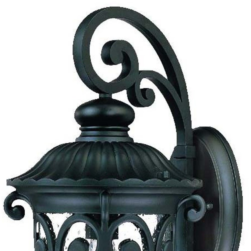 Zoomed in view of the wall plate, scroll arm, and lantern top of the Naples matte black outdoor wall lantern with ornate scrollwork and seeded glass shade