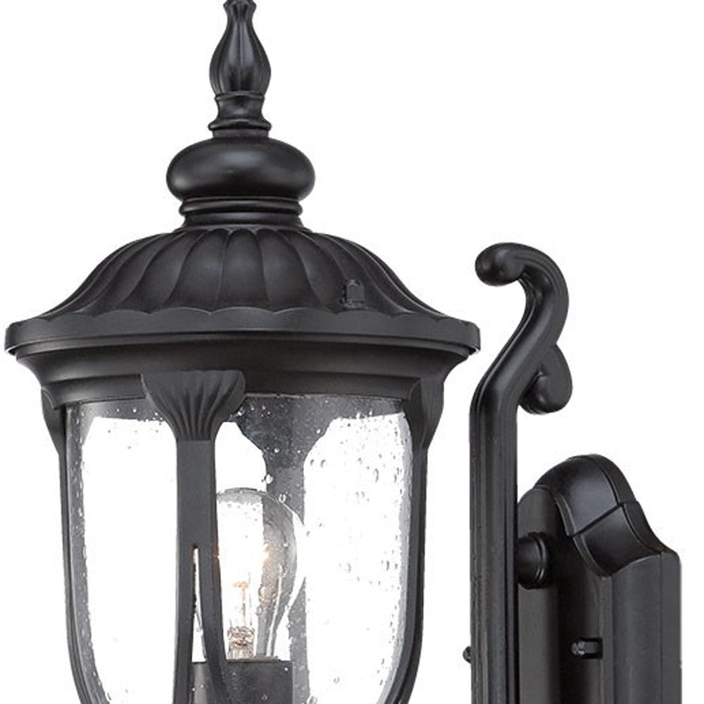 Zoomed in view of the wall plate, arm, and fixture top of the lighted Matte black cast aluminum outdoor glass lantern wall light with clear panels