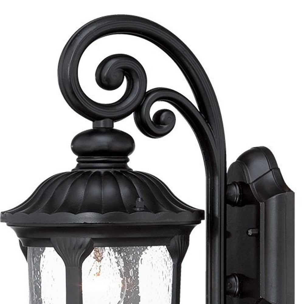Zoomed in view of the wall plate, scroll arm, and lantern top of the lighted Matte black cast aluminum outdoor glass lantern wall sconce with clear panels