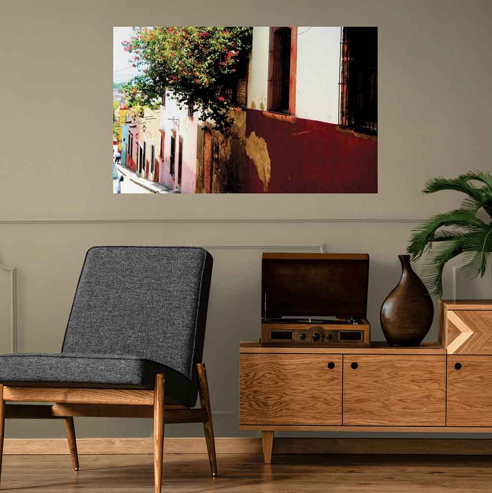 48 inch poster of Mexican homes along a street with red floral buses displayed on wall above a retro chair with credenza