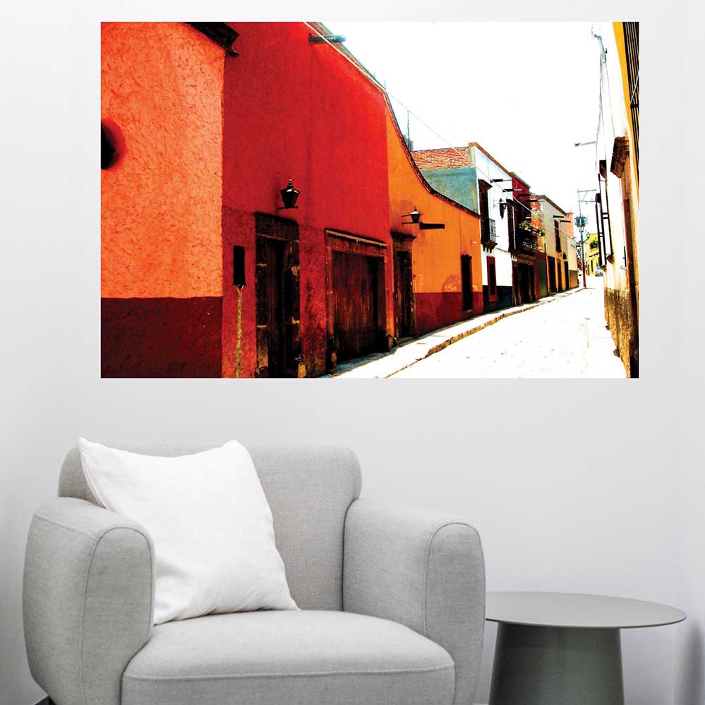 48 inch Colorful Mexican street scene poster with red and orange buildings displayed on a white wall above a gray armchair.