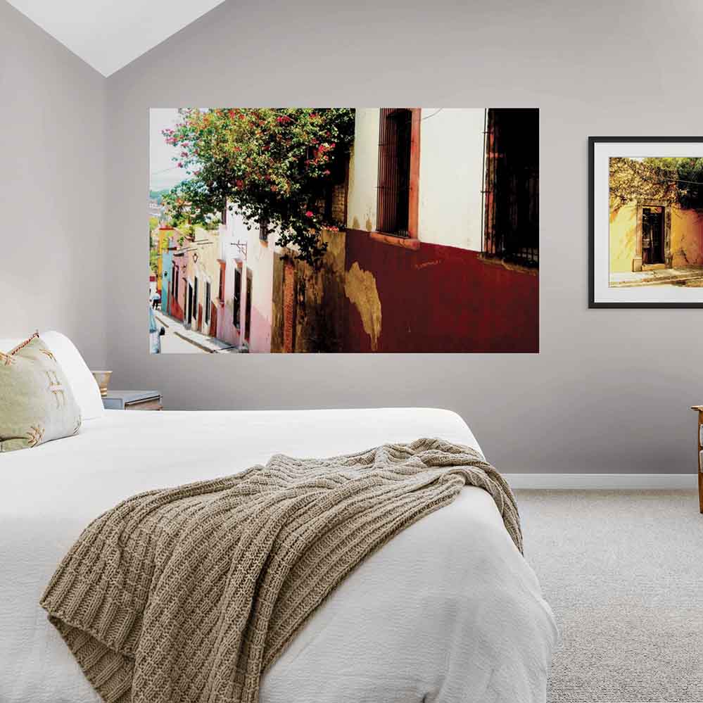 72 inch poster of Mexican homes along a street with red floral buses displayed on wall in a bedroom with bed in the foreground