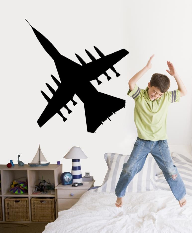 A black vinyl wall decal of a fighter jet is shown on a bedroom wall above a bed, with a young boy jumping excitedly on the bed in the foreground.