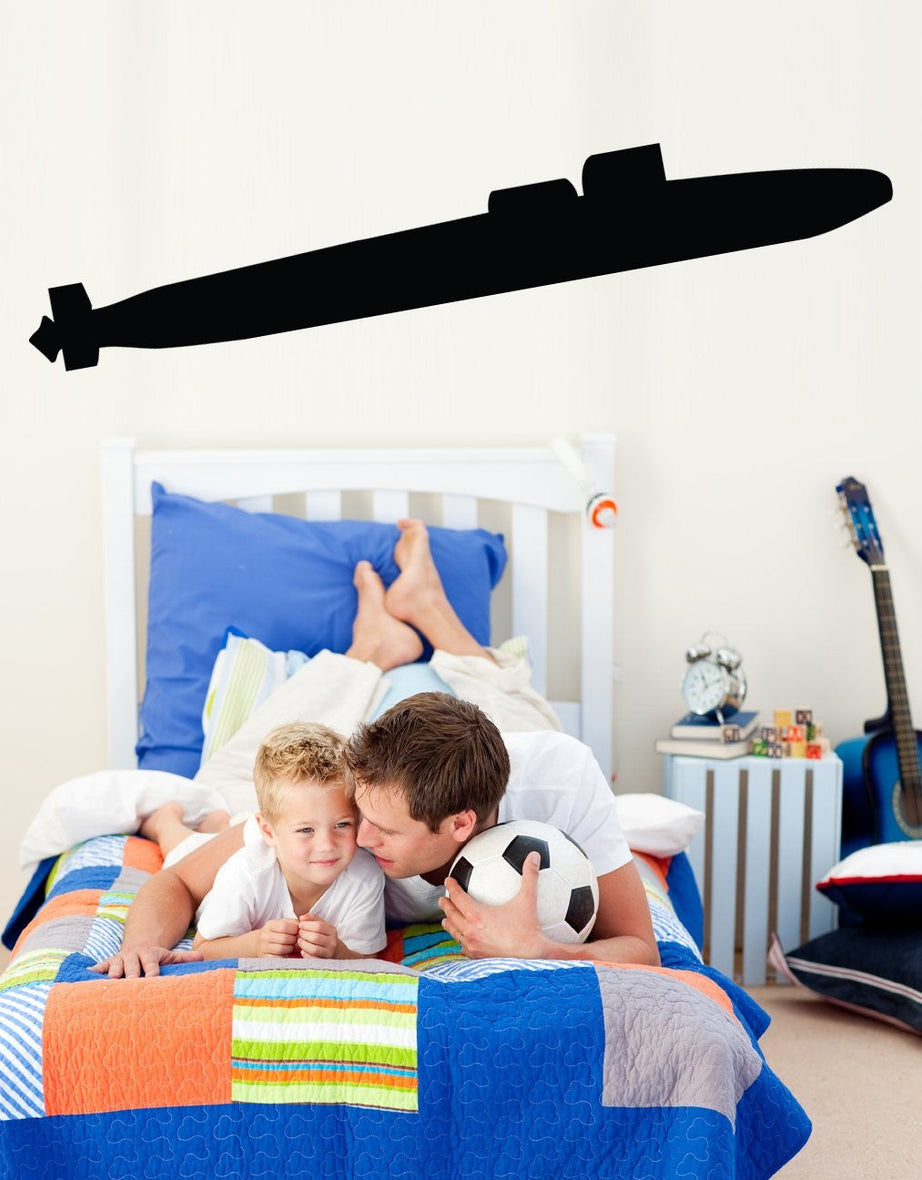 A black submarine wall decal is shown applied to a white wall in a child's bedroom. In the foreground, a father and son are lying on a colorful patchwork bed, and a guitar is visible on a side table in the background.