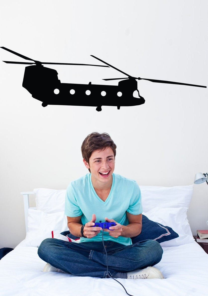 A young man sitting cross-legged on a white bed holding a colorful game controller, with a large black helicopter wall decal displayed above him on a white wall.