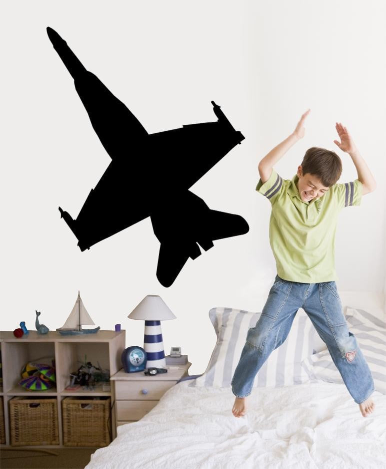 A child jumping excitedly on a bed in a bedroom, with a large black airplane silhouette wall decal displayed on the wall above.