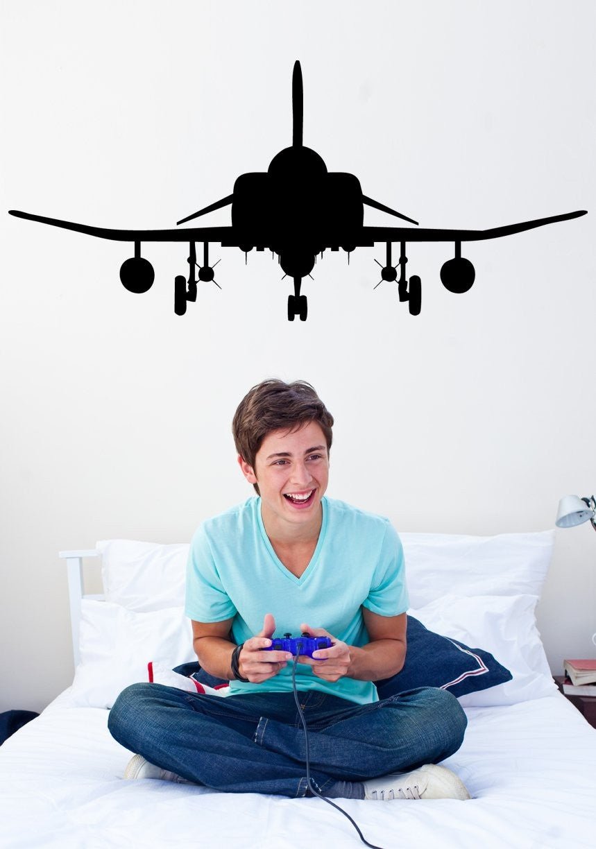 A young man sitting cross-legged on a white bed holding a purple video game controller, with a large black airplane silhouette decal on the wall above his head.