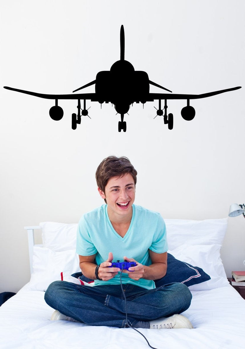 A young man sitting cross-legged on a white bed holding a purple video game controller, with a large black airplane silhouette decal on the wall above his head.