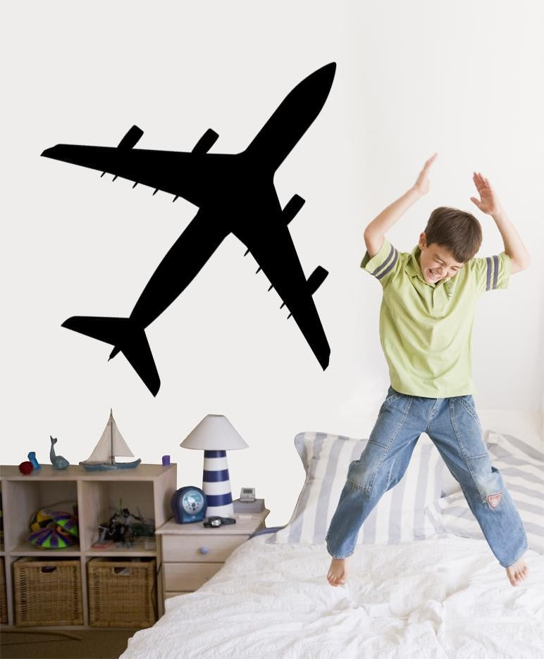 A black airplane wall decal is shown on a bedroom wall above a bed, with a child jumping excitedly on the bed. The room includes a small shelf with toys and a lamp.