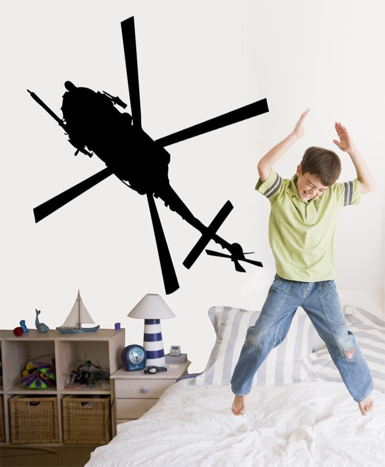 A black vinyl wall decal of a Black Hawk helicopter is shown on a bedroom wall next to a child jumping excitedly on a bed. The room includes a wooden toy shelf with a sailboat model and a striped pillow.