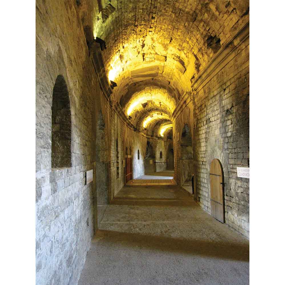Long, narrow stone corridor with arches and a warm glow from ceiling lights in Naimes France Roman Arena wall decal printed | Wallhogs